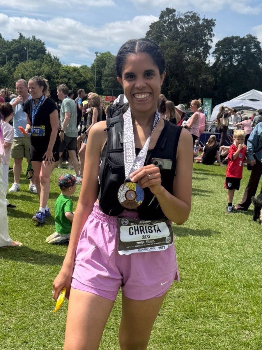 Christa smiling in the sunshine at a race finish, holding up her medal, wearing a race vest and hydration pack with the number 3573 on her bib